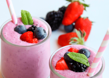 Dos vasos de smoothies con trocitos de fresas y arándanos y decorados con dos sorbetes rosas. En el fondo, difuminados, se ven algunas piezas de fresas y de arándanos.