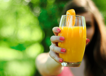 Una chica, con las uñas pintadas de rosa, sujeta un vaso transparente con zumo de naranja.