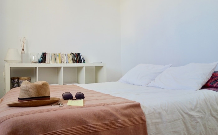 cama en habitación minimalista, con colcha rosada, sombrero, llaves y gafas de sol sobre la cama y estantería blanca al fondo