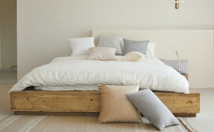 cama sin cabecero, sábana blanca y cojines en azules, grises y rosados, con somier de madera