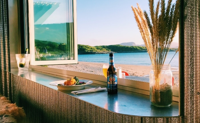 ventana con vistas a la playa con balda a modo de mesa, con una cerveza, una planta y un plato de comida