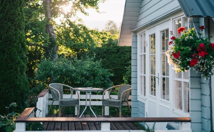 vista de porche con suelo de madera de pequeña casa en blanco y azul con buen jardín. Hay una mesa redonda con dos sillas de mimbre