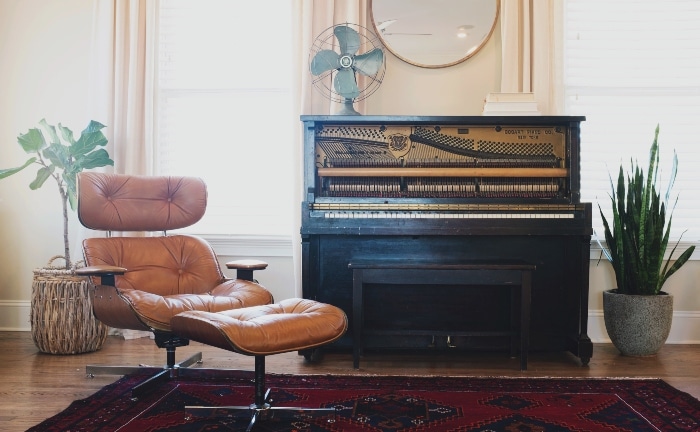 butaca de salita junto a un piano con un ventilador y un espejo redondo en la pared, con alfombra y dos plantas en el suelo
