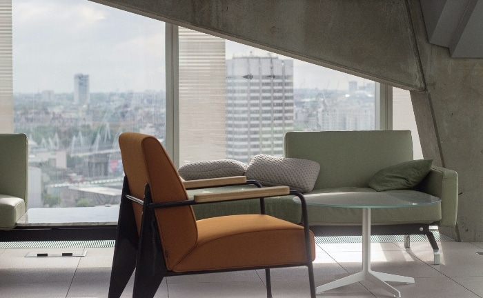 salón con gran ventanal, sofá verde con cojón beige, silla anaranjada con metal y mesa blanca de cristal de centro
