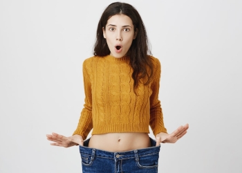 Portrait of attractive slim woman expressing astonishment and amazement, stretching jeans, showing she lost weight after signing on fitness, standing over gray background. Visible result of exercises.