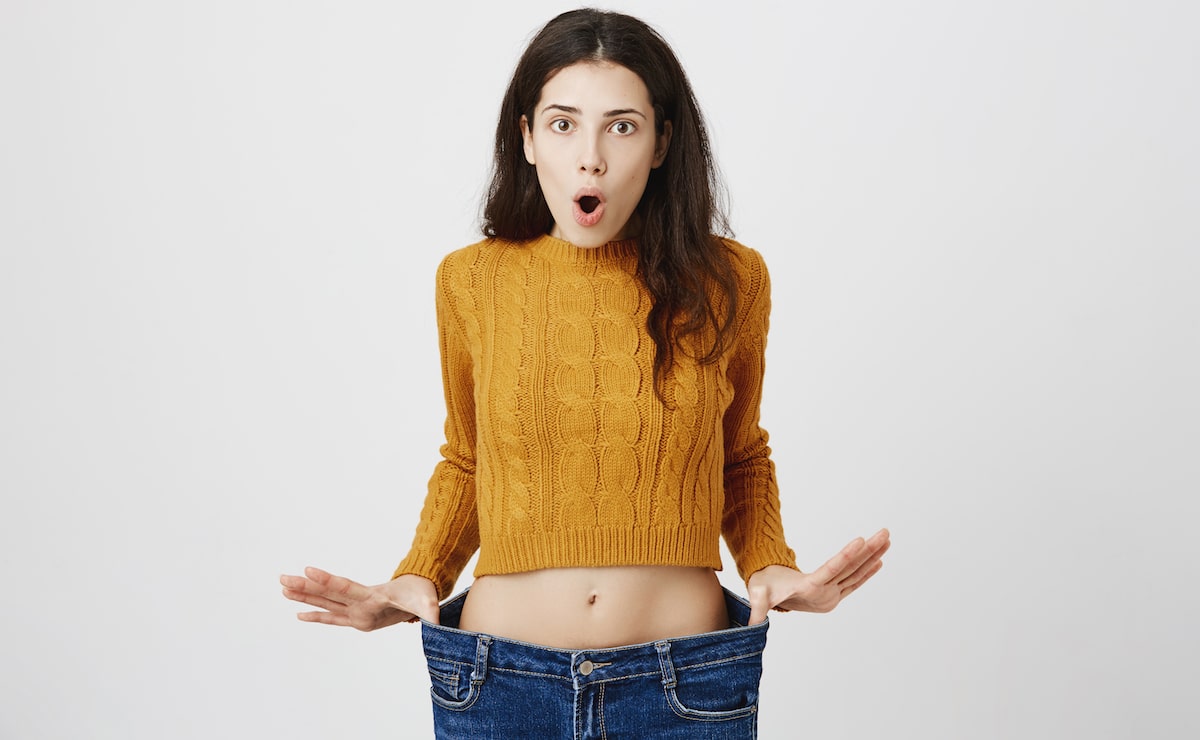 Portrait of attractive slim woman expressing astonishment and amazement, stretching jeans, showing she lost weight after signing on fitness, standing over gray background. Visible result of exercises.